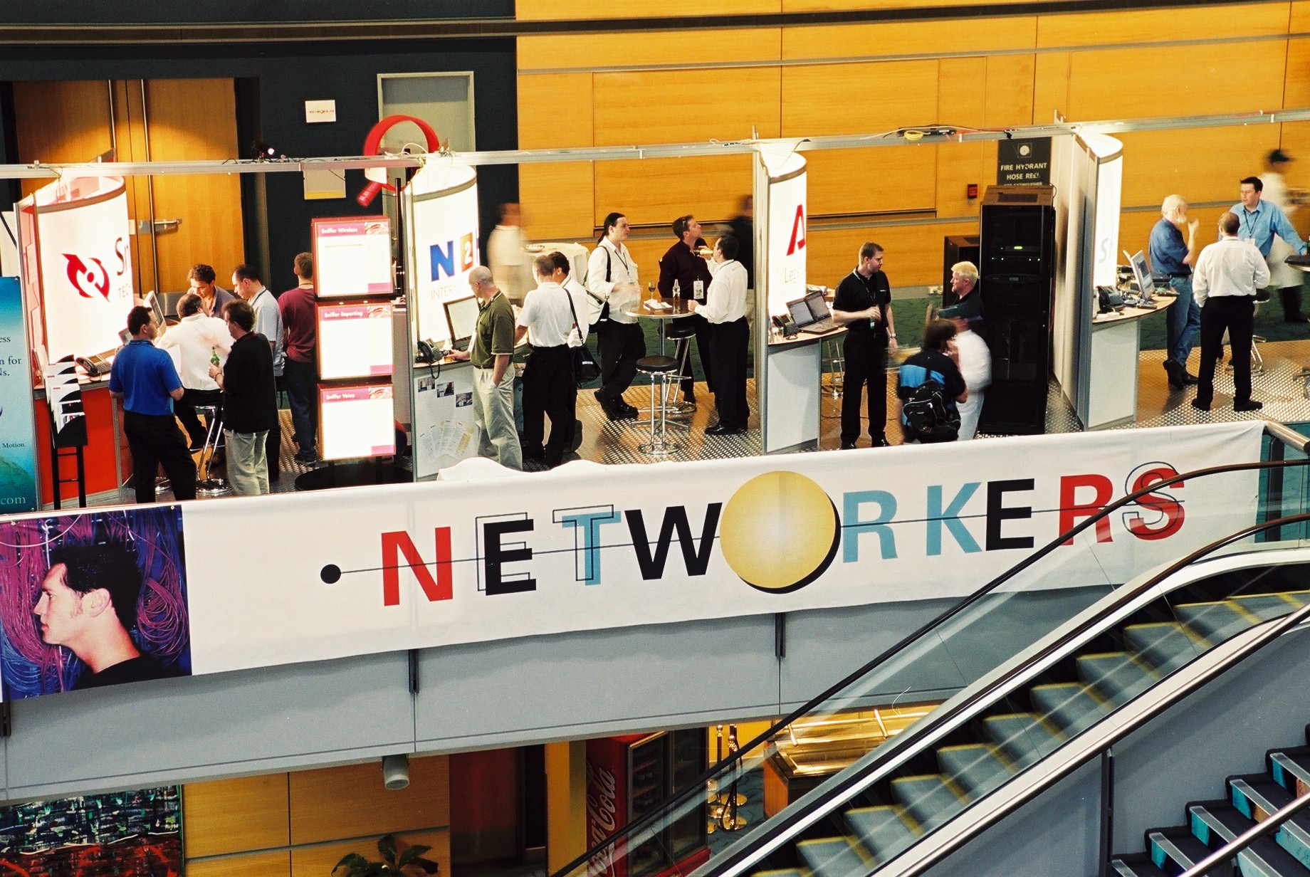 A banner reads "networkers" in funky font as conference attendees mill between booths at Networkers