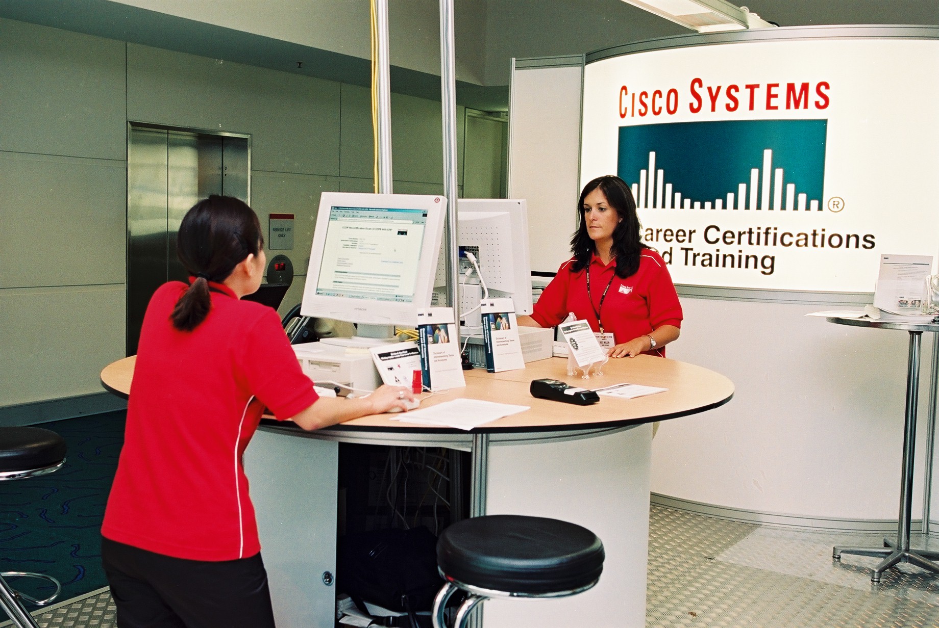 Two cisco representatives stand at computer terminals in front of the Cisco Systems Career Certification and Training booth at Networkers