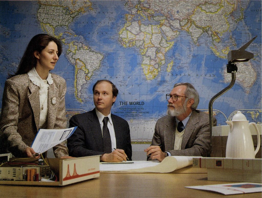 Two men, Len Bosack in a black suit and an unidentified man in tweed, sit at a table looking up at Sandy Lerner, a woman in a business suit standing in profile holding documents.